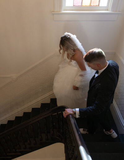 A bride and groom ascend a staircase with a stained glass window above them. The groom is dressed in a suit, and the bride is in a white gown.