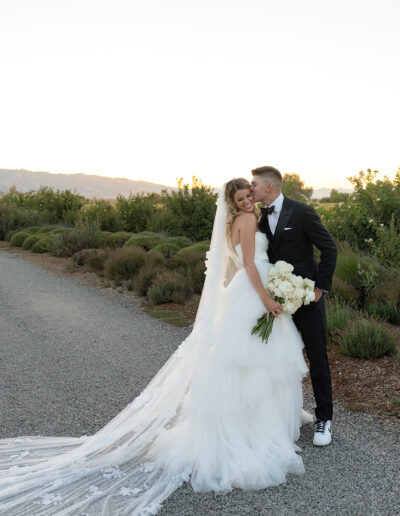 A bride and groom stand on a gravel path, with the groom kissing the bride's cheek. She holds a bouquet of white flowers, and they are surrounded by greenery at sunset.