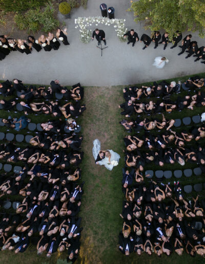 Aerial view of an outdoor wedding ceremony with guests seated in rows on either side of the aisle, where a couple stands in front of an officiant.