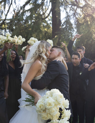 A bride and groom kiss outdoors, surrounded by bridesmaids in black dresses holding flowers and groomsmen in black suits, with trees and sunlight in the background.