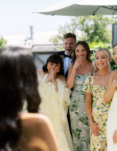 A group of five people in formal attire stand under an umbrella, smiling and reacting joyfully towards a person in the foreground.