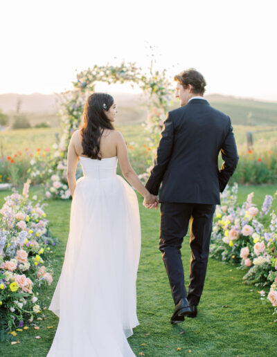 A bride and groom hold hands, walking on grass toward a floral arch. The bride wears a white dress, and the groom is in a dark suit. Flowers line their path, with a scenic backdrop.