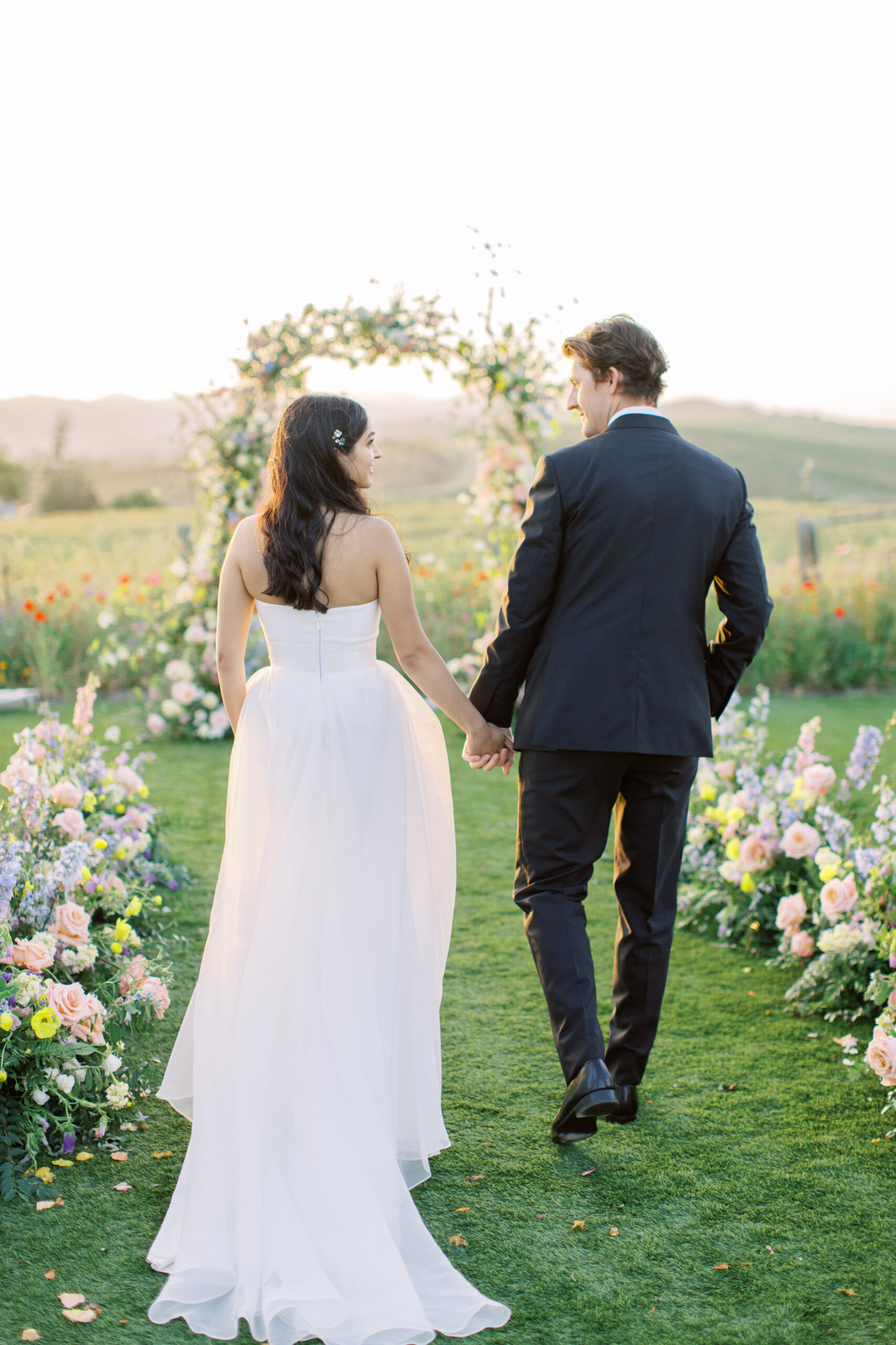 A bride and groom hold hands, walking on grass toward a floral arch. The bride wears a white dress, and the groom is in a dark suit. Flowers line their path, with a scenic backdrop.