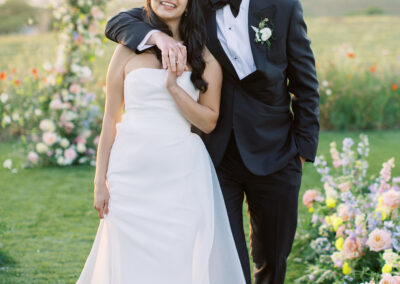 A bride and groom stand together outdoors in formal wedding attire, with a floral arch and scenic landscape in the background.