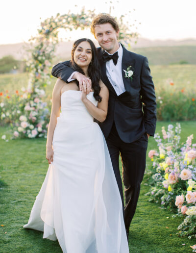 A bride and groom stand together outdoors in formal wedding attire, with a floral arch and scenic landscape in the background.