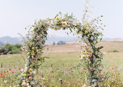 A floral arch made of pastel flowers stands on a grassy field with a backdrop of rolling hills and flowers.