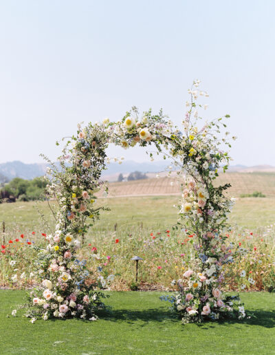 A floral arch made of pastel flowers stands on a grassy field with a backdrop of rolling hills and flowers.