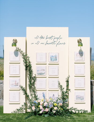 Elegant seating chart display with yellow panels, floral arrangements, and calligraphy reading "all the best people in all our favorite places." Cards are arranged in a grid.