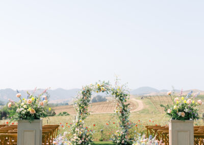 Outdoor wedding setup with a floral arch, wooden chairs, and decorative flower arrangements on a green lawn, with a scenic landscape in the background.