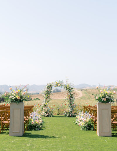 Outdoor wedding setup with a floral arch, wooden chairs, and decorative flower arrangements on a green lawn, with a scenic landscape in the background.