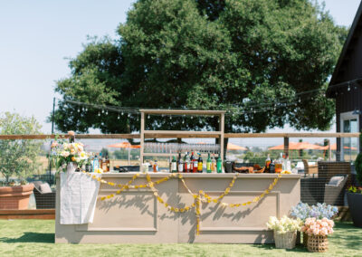 Outdoor bar setup with various bottles and decorations, including a garland and flowers. Large tree in the background, sunny weather.