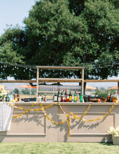 Outdoor bar setup with various bottles and decorations, including a garland and flowers. Large tree in the background, sunny weather.