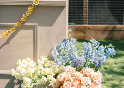 Bar setup outdoors with bottles and citrus garnish on a counter. Baskets of pink roses, white flowers, and blue flowers are placed on the grass below.