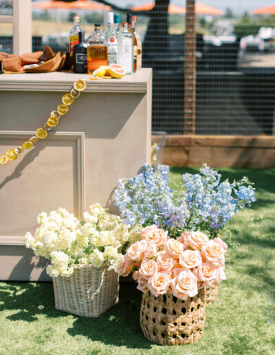 Bar setup outdoors with bottles and citrus garnish on a counter. Baskets of pink roses, white flowers, and blue flowers are placed on the grass below.