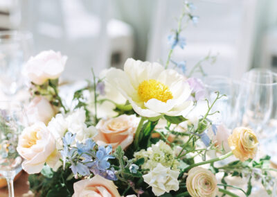 Floral centerpiece with white and pink flowers, including a prominent white peony and scattered greenery, on a table with glassware in the background.