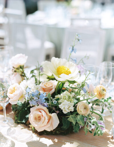 Floral centerpiece with white and pink flowers, including a prominent white peony and scattered greenery, on a table with glassware in the background.