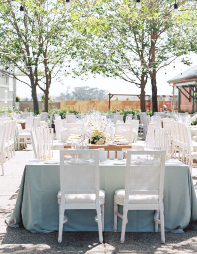 Outdoor wedding reception setup with white chairs, round tables covered with light blue tablecloths, decorated with floral centerpieces, and string lights overhead.