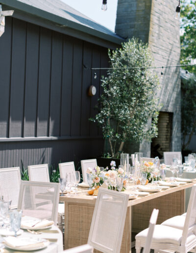 Outdoor dining setup with beige chairs around wooden tables. The tables are decorated with flowers and candles. A gray building and string lights are in the background.