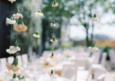 Hanging white and peach flowers on thin strings in focus; blurred outdoor setting with white chairs and tables in the background.