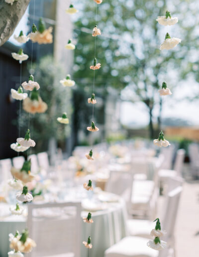 Hanging white and peach flowers on thin strings in focus; blurred outdoor setting with white chairs and tables in the background.