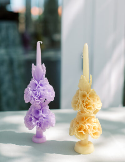 Two ornate candles, one purple and one yellow, with floral designs, stand on a table in sunlight.