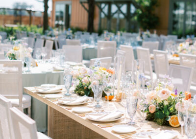 Outdoor wedding reception with elegantly set tables, decorated with floral centerpieces and glassware, arranged in a garden setting.