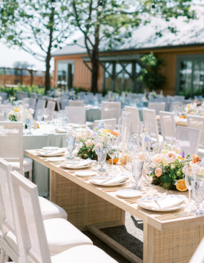 Outdoor wedding reception with elegantly set tables, decorated with floral centerpieces and glassware, arranged in a garden setting.