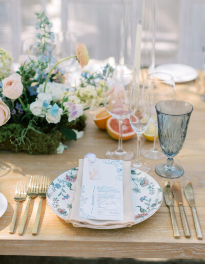 Elegant table setting with floral-patterned plate, gold cutlery, crystal glasses, and a floral centerpiece. A menu lies on the plate beside citrus fruits.