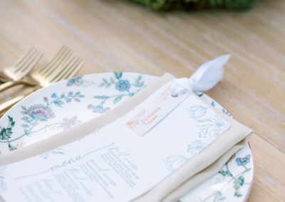 A table setting with a floral-patterned plate, a folded menu, gold cutlery, and a floral centerpiece.