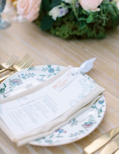 A table setting with a floral-patterned plate, a folded menu, gold cutlery, and a floral centerpiece.