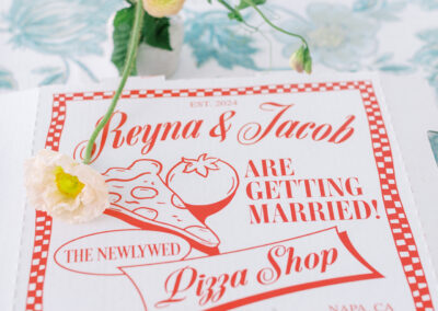 Pizza box with text: "Reyna & Jacob are getting married! The Newlywed Pizza Shop, 05.18.2024, Napa, CA," decorated with flowers and a floral tablecloth.