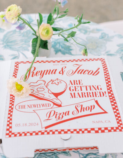 Pizza box with text: "Reyna & Jacob are getting married! The Newlywed Pizza Shop, 05.18.2024, Napa, CA," decorated with flowers and a floral tablecloth.