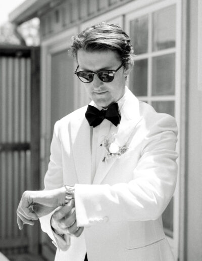 A man in a white tuxedo and sunglasses checks his watch outside near a building.