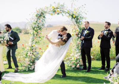 A bride and groom kiss in front of a floral arch during an outdoor wedding ceremony, surrounded by people clapping. The scene is bright and cheerful with a clear sky.