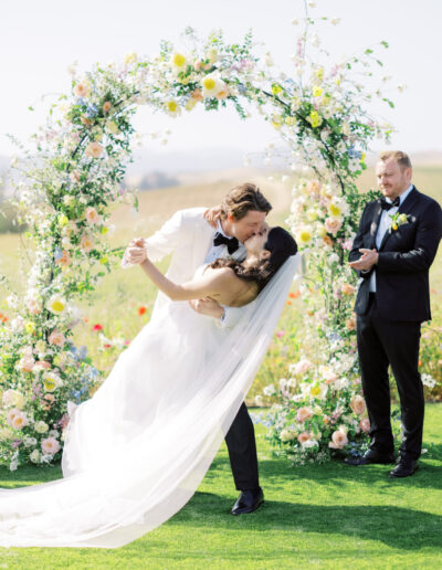 A bride and groom kiss in front of a floral arch during an outdoor wedding ceremony, surrounded by people clapping. The scene is bright and cheerful with a clear sky.