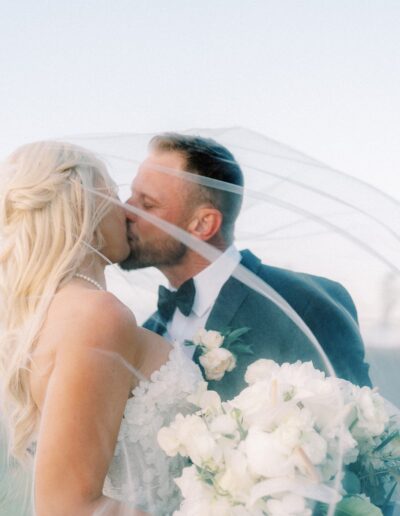 A bride and groom are kissing under a veil, holding a bouquet of white flowers.