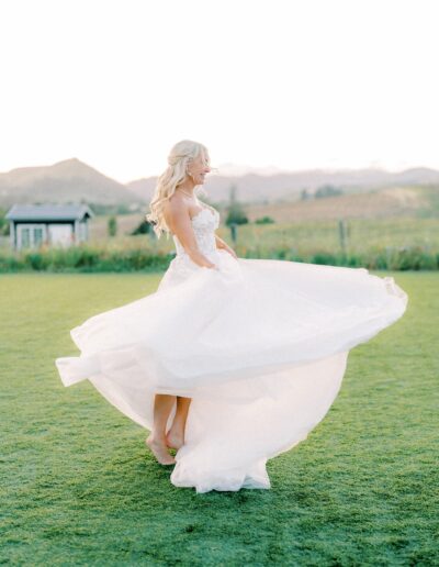 A bride in a white dress twirls on a grassy field with mountains and a small building in the background.