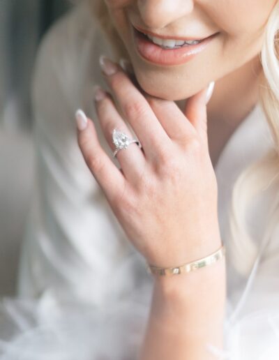 A person with long, blonde hair smiles while resting their chin on their hand, displaying a ring and a bracelet.