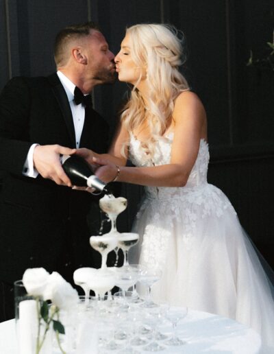 A couple in formal attire kisses while pouring champagne into a tower of glasses.