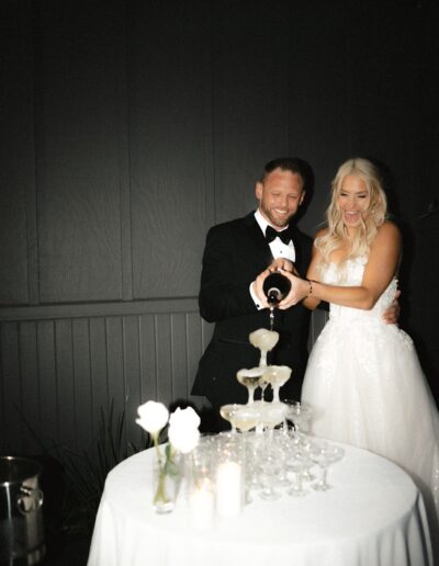 A couple in formal attire is pouring champagne into a tower of glasses on a table with candles and white roses.