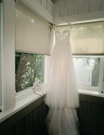 A white wedding dress hangs on a window frame beside a pair of white heels in a sunlit room.