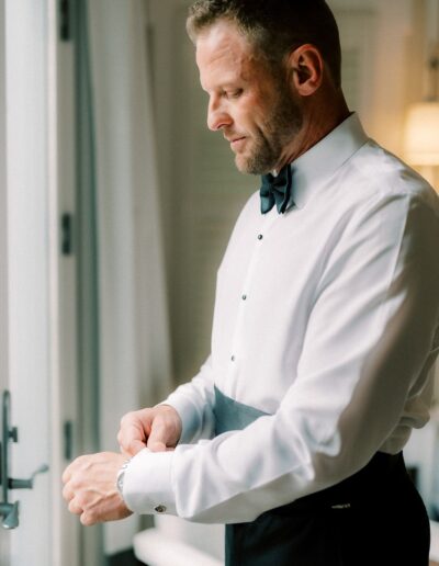 A man in formal attire adjusts his cufflink near a window, wearing a white shirt with a black bow tie and cummerbund.