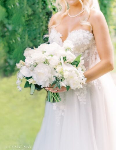 Bride in a white lace gown holding a bouquet of white flowers, standing outdoors with greenery in the background.