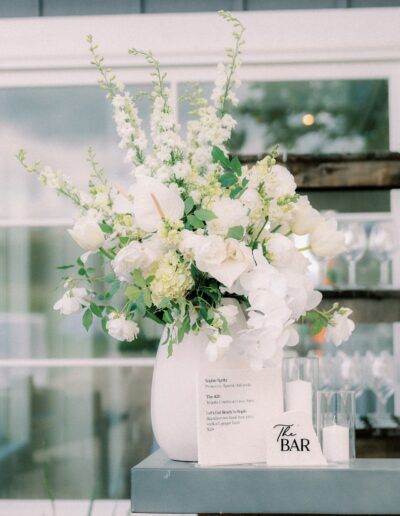 White floral arrangement in a vase on a table next to a sign labeled "The Bar" and a menu. Glasses are seen in the background.