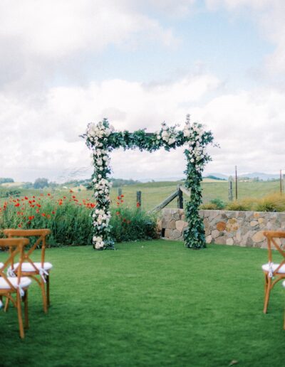 Outdoor wedding ceremony setup with wooden chairs facing a floral arch. Green grass and open sky in the background.