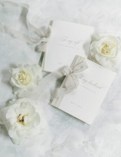 Two white wedding vow booklets with gray ribbons surrounded by white roses on a textured white surface.