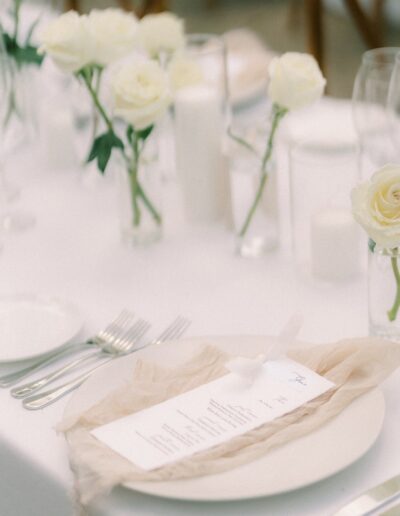 A wedding table setting with white roses, candles, cutlery, and a menu placed on a white plate with a beige napkin.