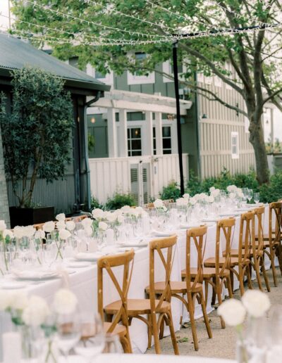 Outdoor dining setup with long tables, white tablecloths, wooden chairs, and floral centerpieces under string lights. Trees and buildings in the background.