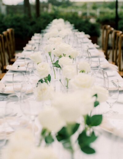 A long dining table is elegantly set with white rose centerpieces, wine glasses, and place settings. The setting is outdoors with blurred greenery in the background.