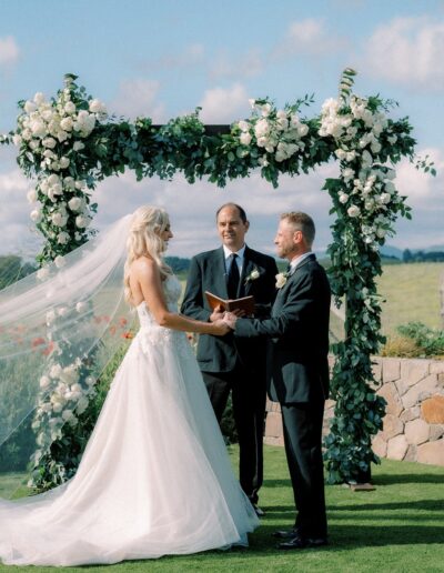 Bride and groom hold hands during an outdoor wedding ceremony under a floral arch, with a person officiating.